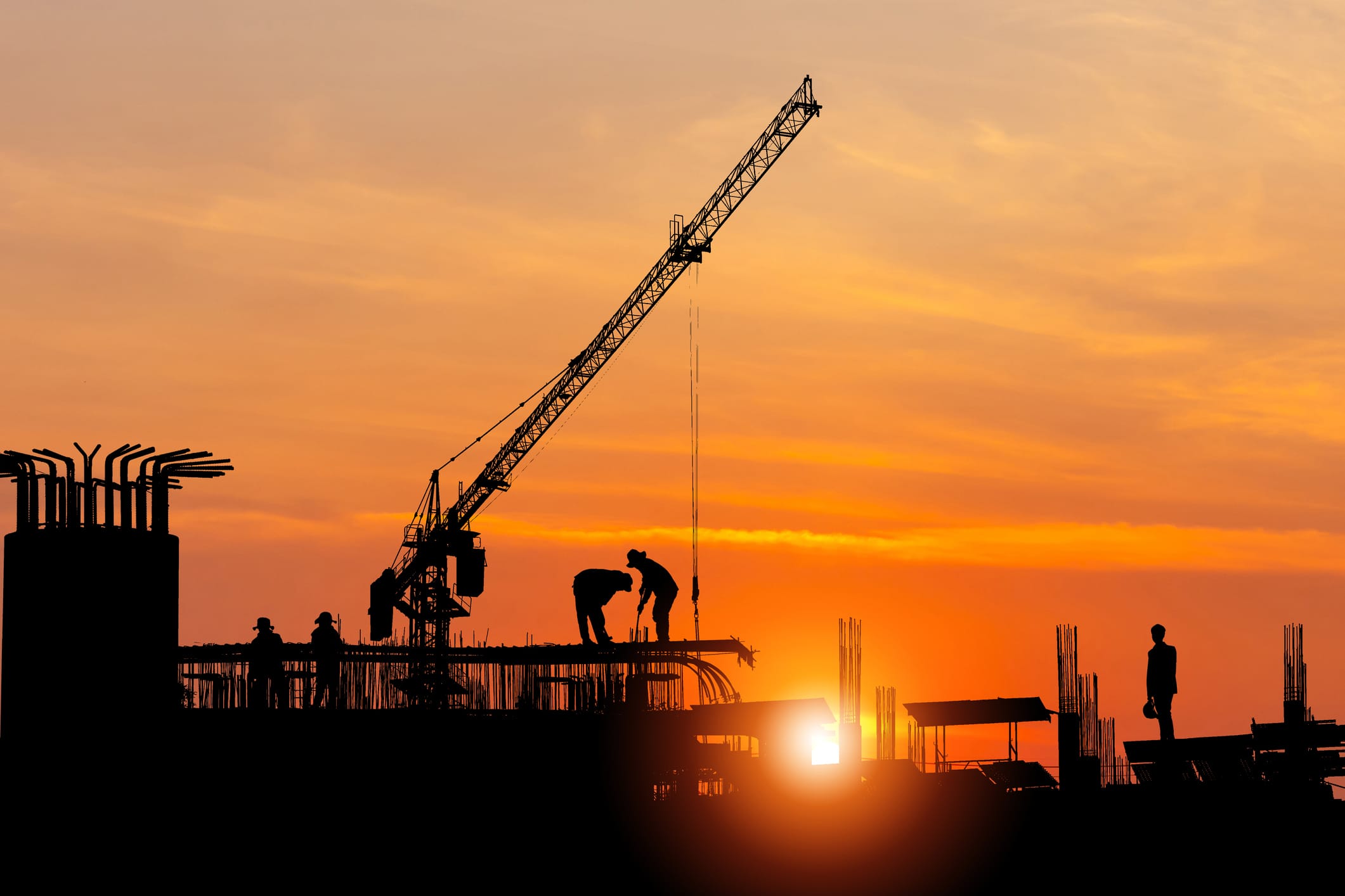 The Silhouette Of A Construction Crane, Building, And Workers Set In Front Of A Beautiful Orange Sunset.